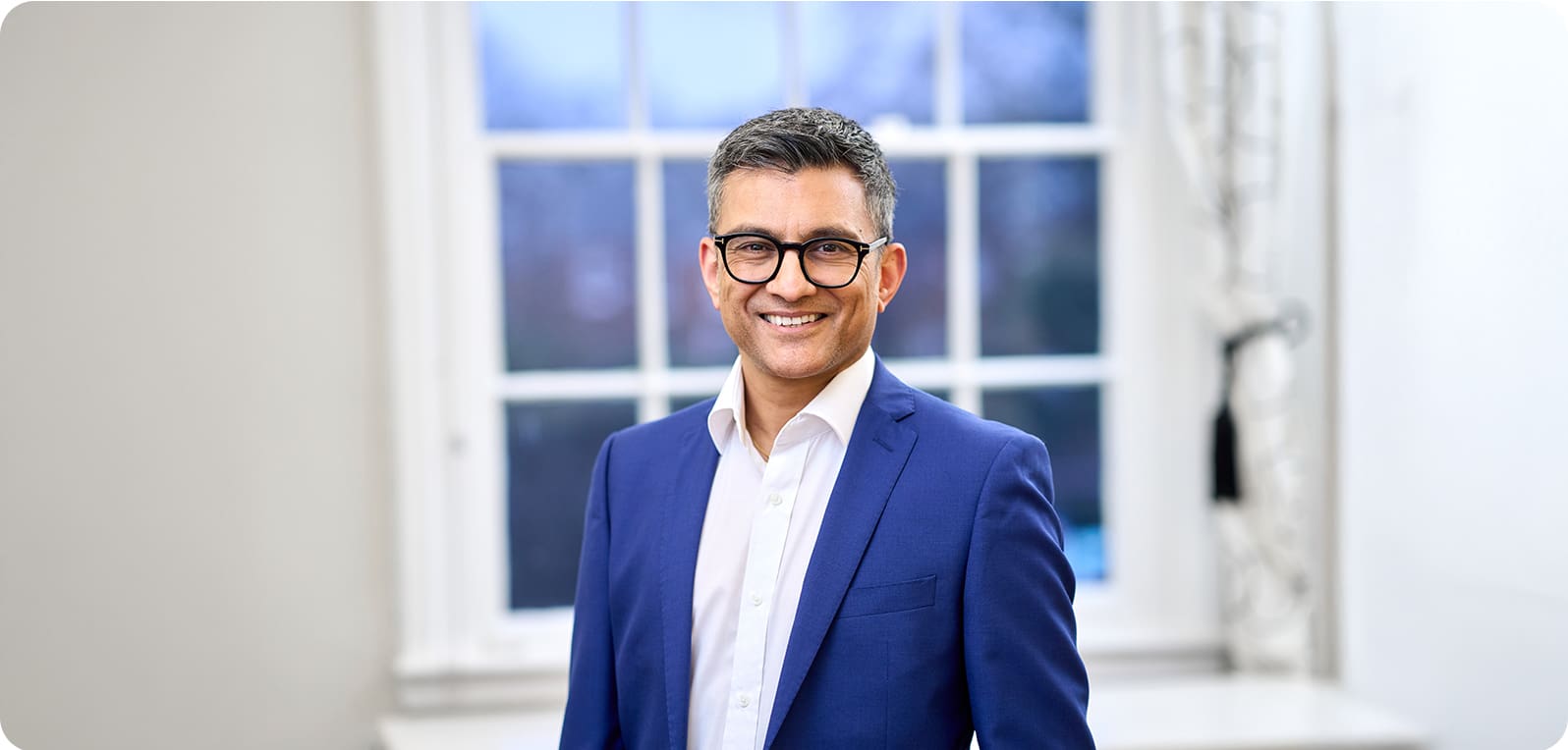 Dr. Nick Makwana in a blue suit and white shirt stands in a bright room with a large window in the background, conveying a professional and welcoming atmosphere.