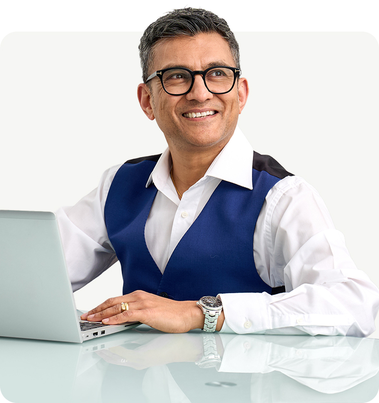 Dr. Nick Makwana in glasses and a blue vest smiles while working on a laptop. He exudes confidence and professionalism against a plain white background.