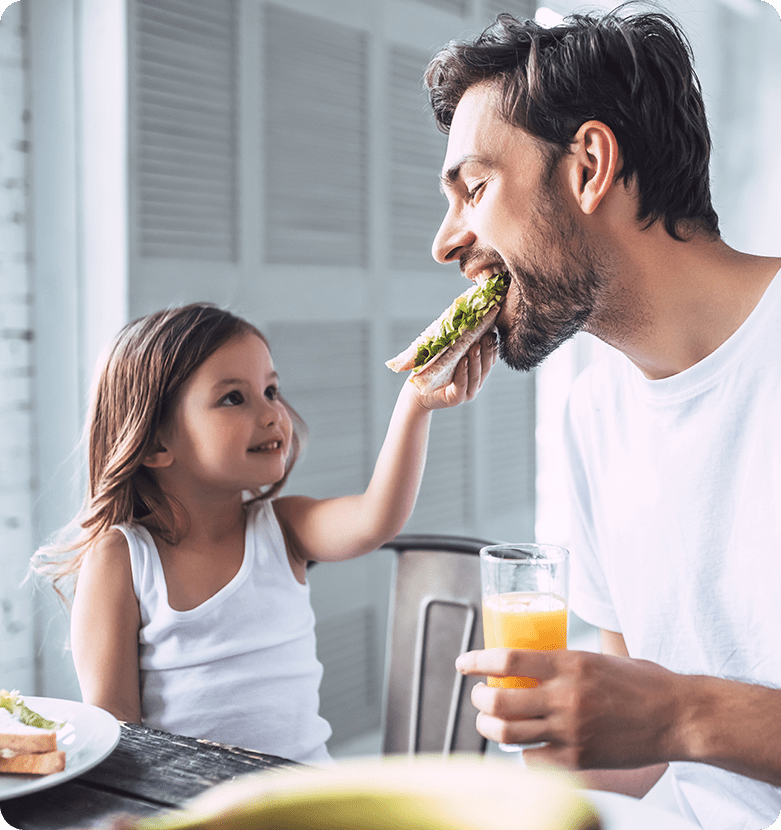 A smiling child feeds her dad a sandwich as he holds a glass of orange juice. They are sitting at a table, sharing a joyful moment in a bright room.