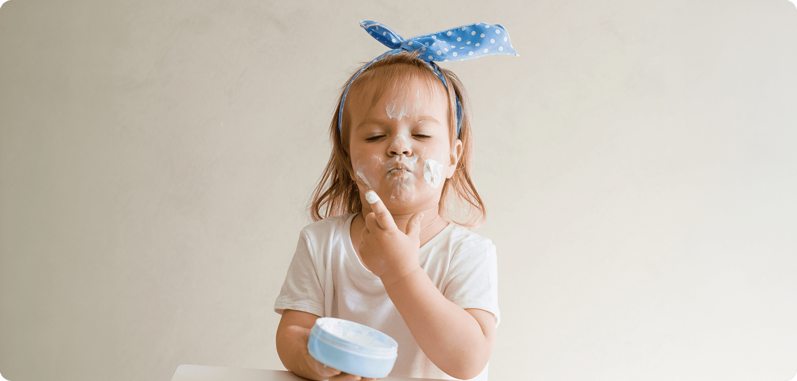 A toddler with a blue polka dot headband applies cream to her face, eyes closed in concentration. She holds a cream, expressing playful curiosity.