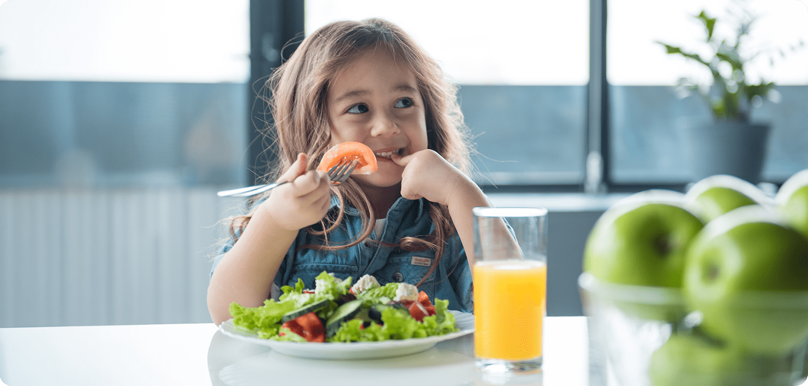 A young girl joyfully eats a salad, holding a fork with a tomato slice. A glass of orange juice and green apples are on the table. Bright and cheerful scene.