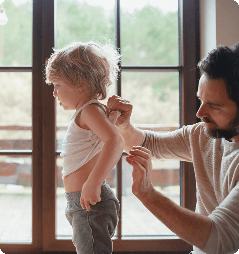 A father helps his young child put on a white tank top near a large window with a scenic view. The scene conveys warmth and care in a cozy home setting.
