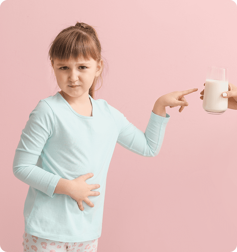 A young girl with light brown hair has a concerned expression as she points at a glass of milk in front of her, holding her other hand to her stomach.