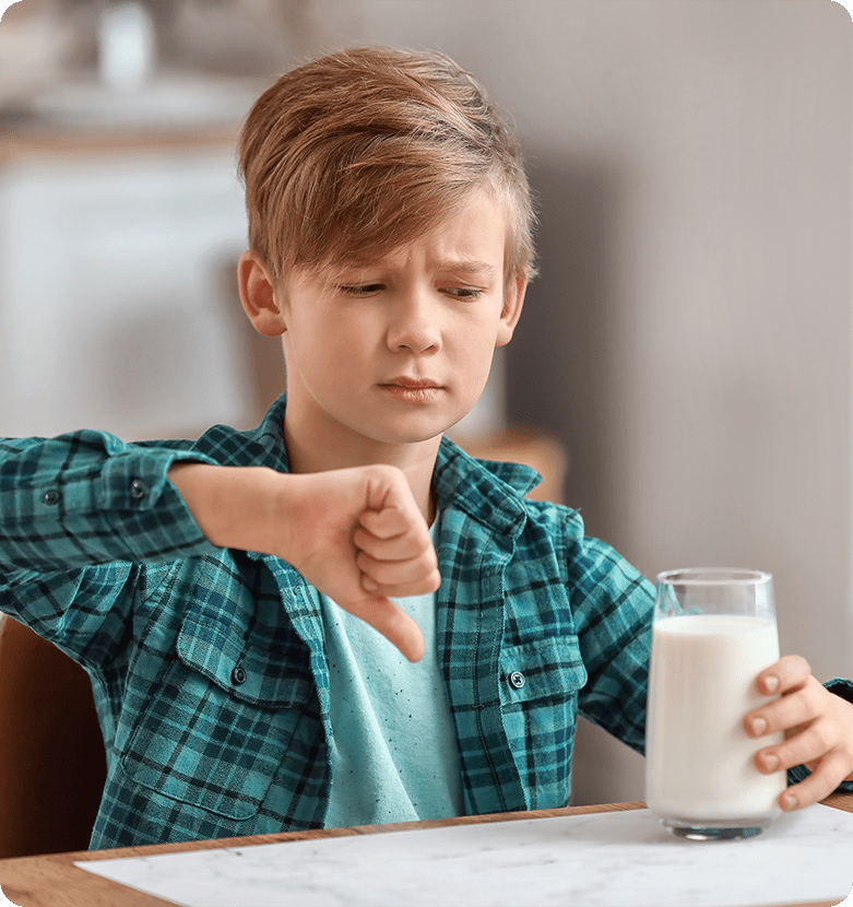 Young boy in a teal plaid shirt frowns and gives a thumbs-down to a glass of milk on a table, expressing dislike or disapproval indoors.