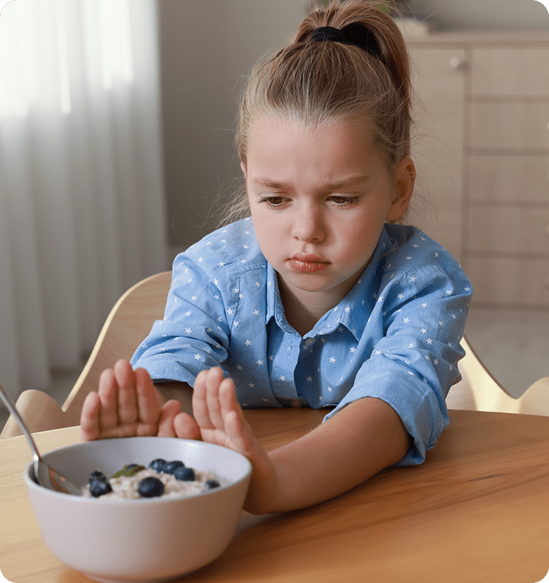 Young girl with a ponytail, wearing a blue shirt, looks disinterested as she pushes a bowl of oatmeal with blueberries away at a wooden table.