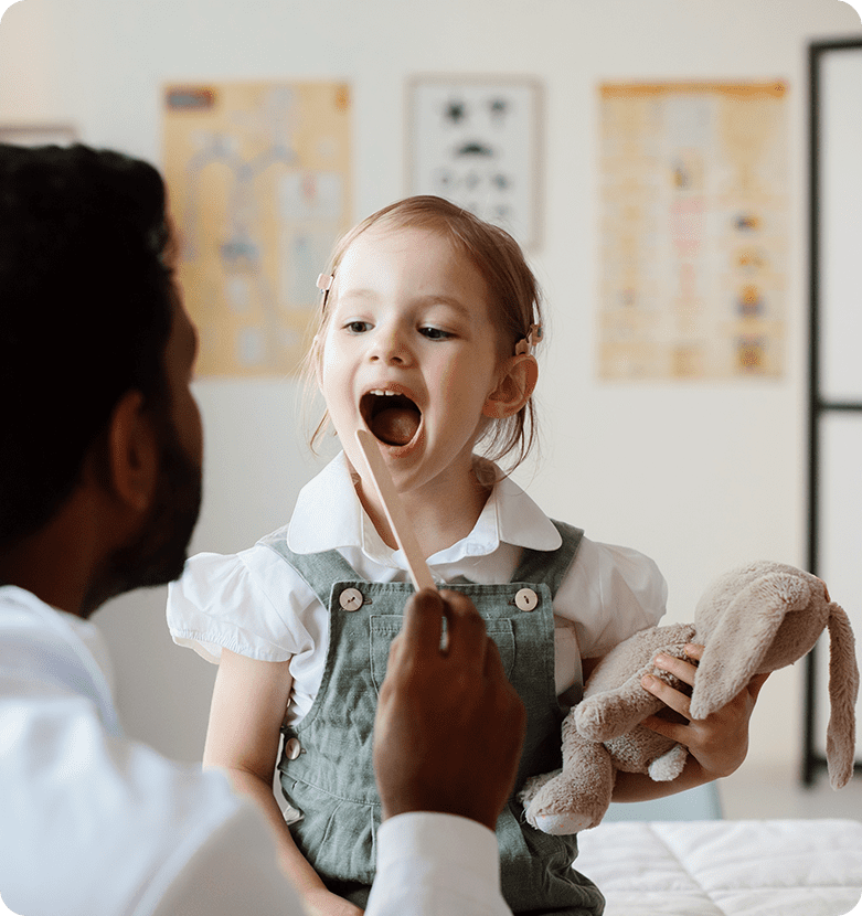 A doctor examines a young girl holding a stuffed rabbit, with a tongue depressor in her mouth. The child appears calm in a clinic setting.