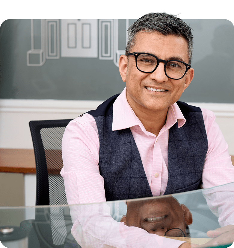 Dr. Nick Makwana, wearing a pink shirt and blue waistcoat, smiles while sitting at a glass table. The background features a chalk-drawn design.