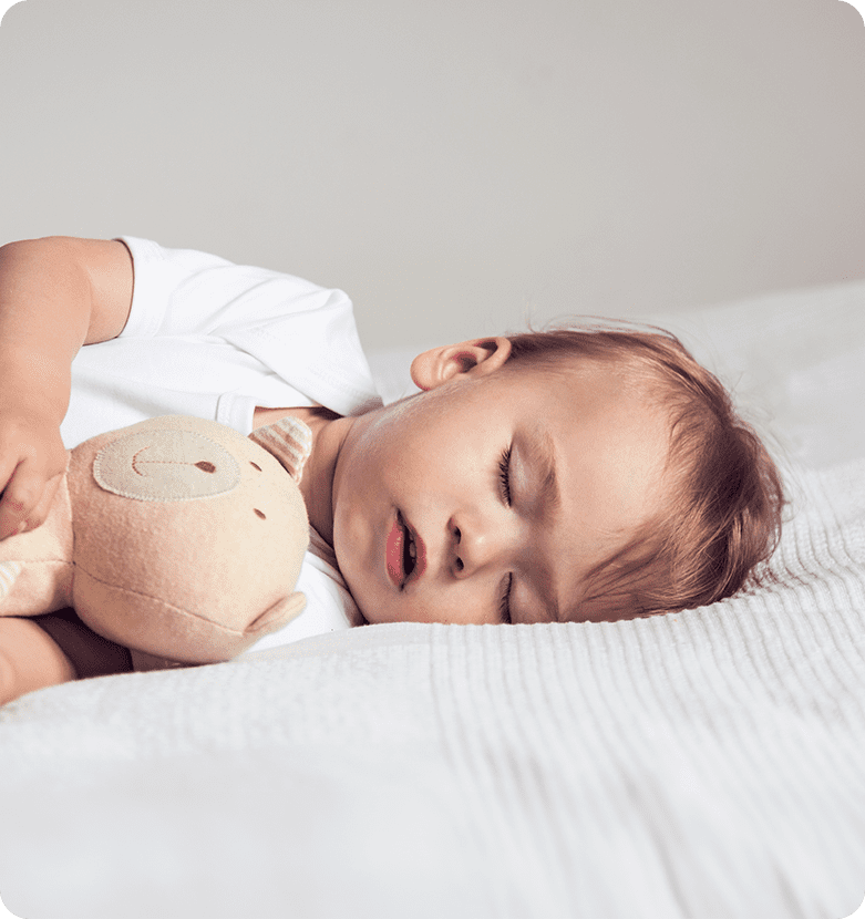 A toddler peacefully sleeps on a white bed, cuddling a soft, beige teddy bear. The scene conveys serenity and comfort in a cozy setting.