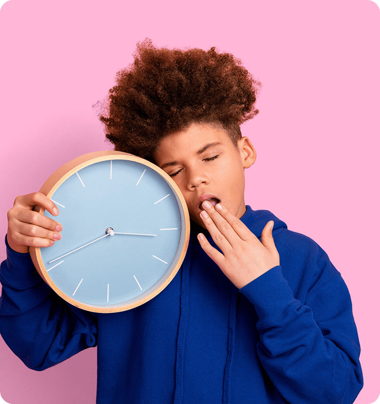 A tired boy in a blue hoodie holds a large wall clock, yawning with eyes closed, against a pastel pink background, conveying sleepiness.
