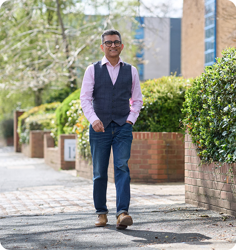 Dr. Nick Makwana walks down a sunlit road lined with green bushes and brick walls, conveying a cheerful, relaxed mood.